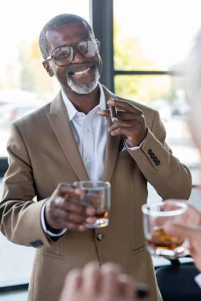 Hombre de negocios afroamericano senior con cigarro y vaso de whisky sonriendo cerca de colega borroso en la oficina - foto de stock