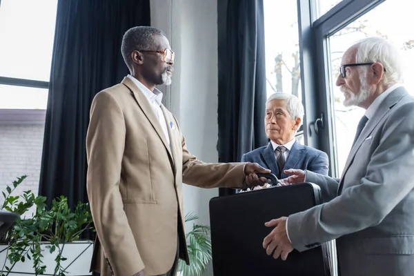 Africano americano uomo d'affari dando valigetta per business partner vicino anziani asiatico collega — Foto stock