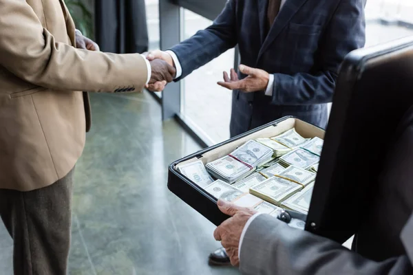 Vista parcial de colegas de negocios interracial estrechando la mano cerca de un hombre sosteniendo el maletín con dinero - foto de stock