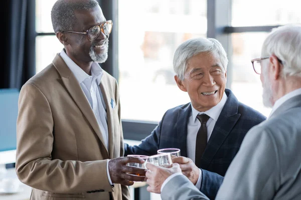 Cheerful multiethnic business colleagues clinking glasses of whiskey in office — Stock Photo