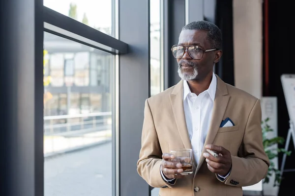 Homme d'affaires afro-américain senior avec cigare et verre de whisky regardant par la fenêtre dans le bureau — Photo de stock