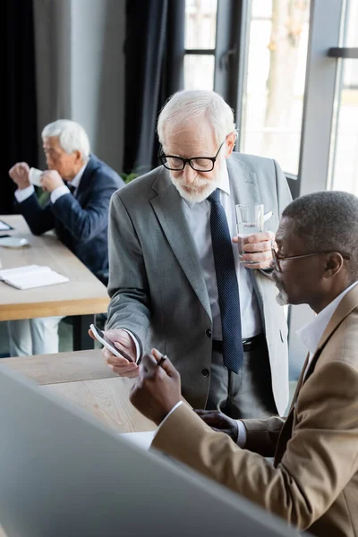 Senior businessman showing smartphone to african american colleague in office — Stock Photo