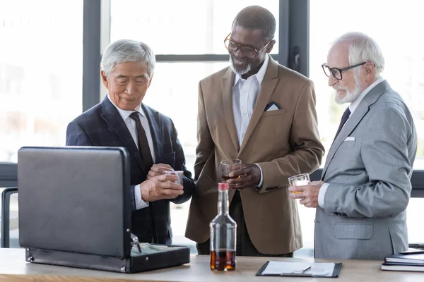 Ancianos hombres de negocios multiétnicos con vasos de whisky sonriendo cerca del maletín y contrato en el escritorio - foto de stock