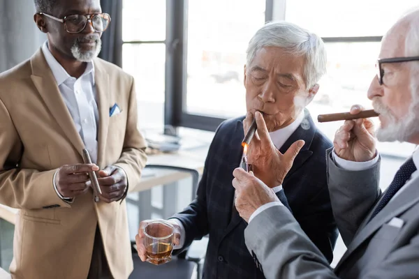 Senior businessman lighting cigarro de asiático colega cerca africano americano negocios socio - foto de stock