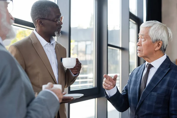 Elderly asian businessman talking to multiethnic colleagues during coffee break — Stock Photo