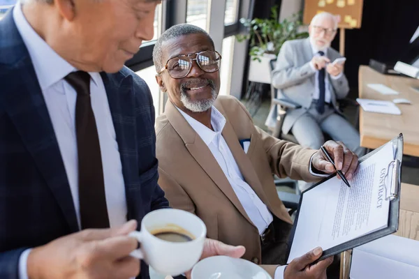 Senior asian businessman with cup of coffee near african american colleague pointing with pen at contract — Stock Photo