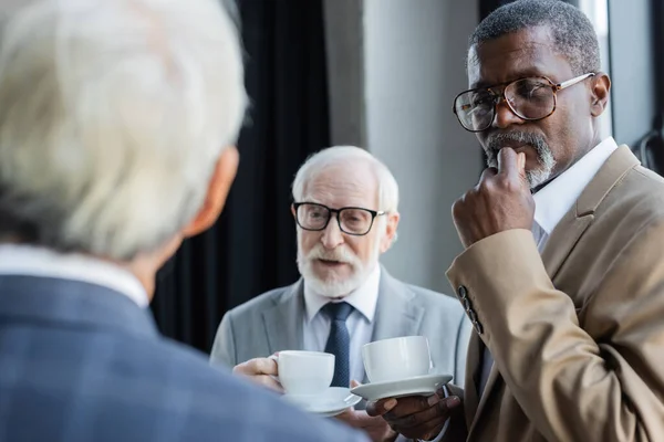 Senior interracial businessmen with coffee cups looking at blurred colleague — Stock Photo