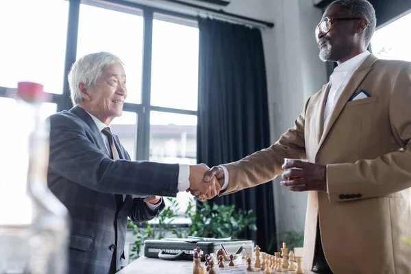 Socios comerciales interraciales de alto nivel estrechando las manos cerca del tablero de ajedrez, borrosa primer plano - foto de stock