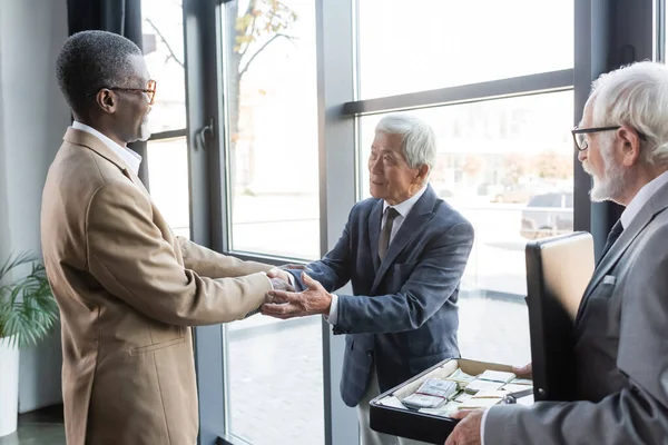 Älterer Geschäftsmann hält Aktentasche mit Geld in der Hand, während interrassische Geschäftspartner im Büro Hände schütteln — Stockfoto
