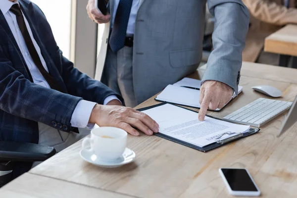 Обрезанный взгляд старшего бизнесмена указывая на контракт рядом с коллегой и размытый кофе чашку — стоковое фото