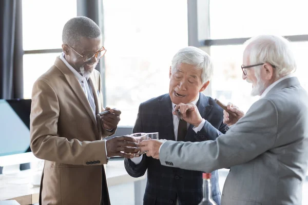 Aufgeregte multiethnische Geschäftspartner mit Zigarren, die im Büro an Gläsern Whiskey klirren — Stockfoto