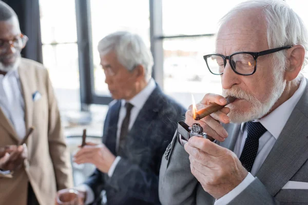 Senior exitoso hombre de negocios iluminación cigarro cerca borrosa socios de negocios multiétnicos hablando en la oficina - foto de stock