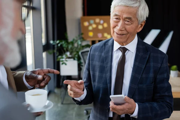 Senior asiatico uomo d'affari con smartphone sorridente vicino sfocato multietnico colleghi — Foto stock