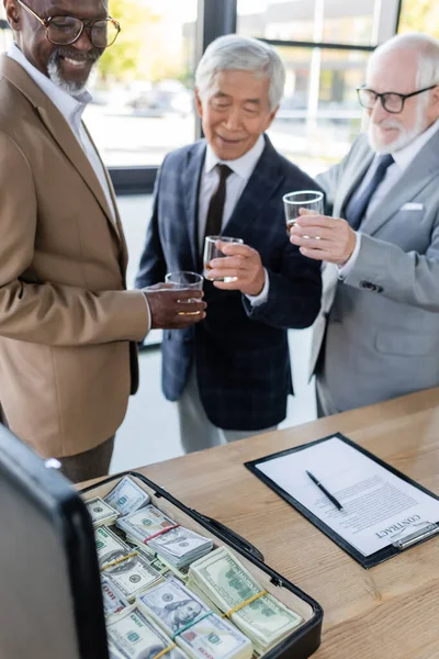 Homens de negócios multiétnicos felizes com copos de uísque perto de contrato e pasta com dinheiro — Fotografia de Stock