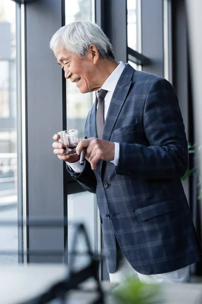 Sênior asiático empresário com uísque e charuto sorrindo enquanto olha para baixo através da janela do escritório — Fotografia de Stock
