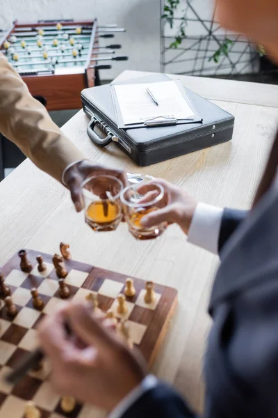 Cropped view of blurred business partners clinking glasses near chessboard and contract on briefcase — Stock Photo