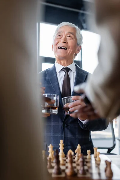 Sênior asiático empresário sorrindo perto de tabuleiro de xadrez e colegas borrados com copos de uísque — Fotografia de Stock