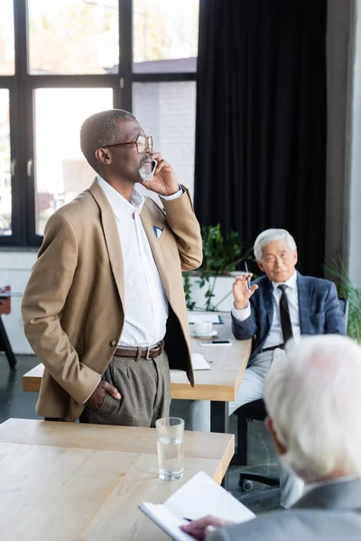 Homme d'affaires afro-américain avec la main dans la poche parler sur smartphone près de collègues au bureau — Photo de stock