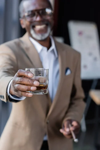 Hombre de negocios afroamericano borroso sonriendo mientras sostiene un vaso de whisky en la mano extendida - foto de stock