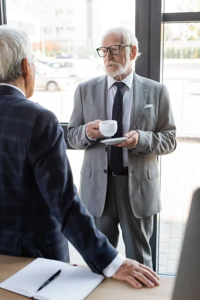 Anziano uomo d'affari in occhiali con in mano una tazza di caffè mentre parla con un collega offuscato — Foto stock