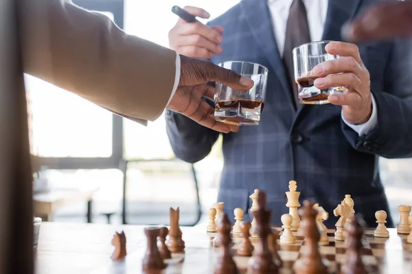 Cropped view of successful interracial business partners holding glasses of whiskey above blurred chessboard — Stock Photo