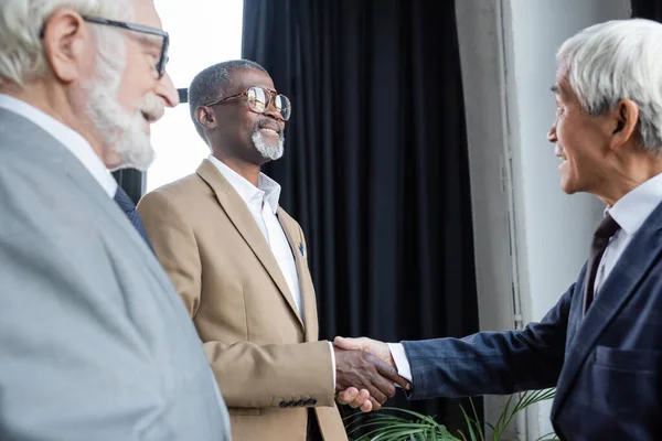 Desfocado sênior empresário sorrindo perto africano americano e asiático parceiros de negócios apertando as mãos no escritório — Fotografia de Stock