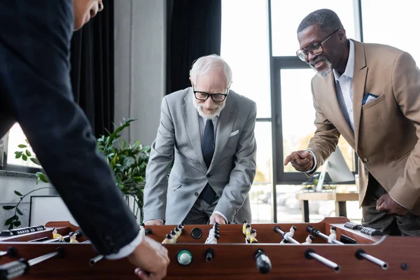 Lächelnder afrikanisch-amerikanischer Geschäftsmann zeigt mit dem Finger, während ältere Kollegen Tischfußball spielen — Stockfoto