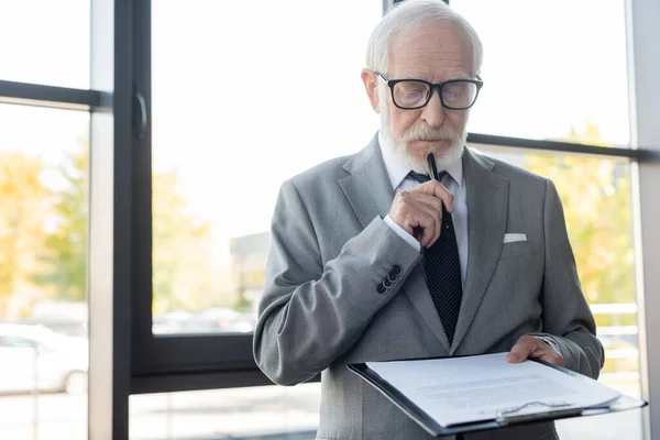 Nachdenklicher Senior-Geschäftsmann hält Stift, während er Vertrag im Amt liest — Stockfoto