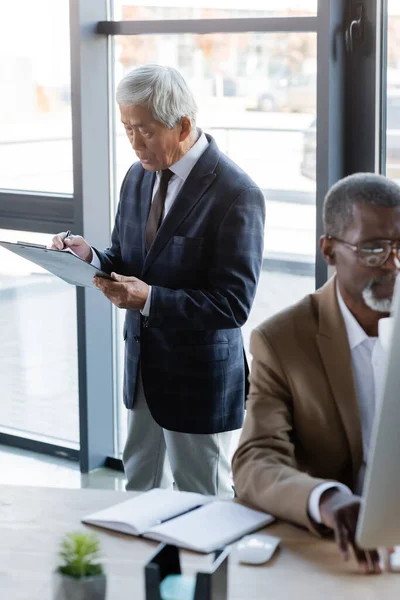 African american businessman working on blurred computer near senior asian colleague with clipboard — Stock Photo