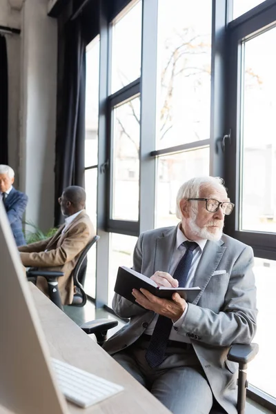 Senior businessman writing in notebook while looking through window in office — Stock Photo