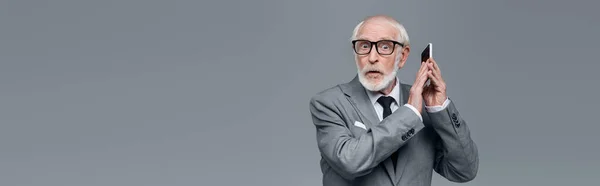 Shocked senior businessman looking at camera while covering cellphone with hand isolated on grey, banner — Stock Photo