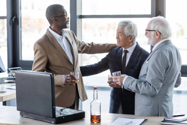 Alegres empresarios multiétnicos con vasos de whisky saludándose cerca del contrato y maletín con dinero - foto de stock