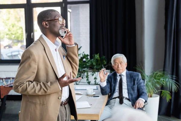 Africano americano empresário falando no celular perto turvo asiático colega — Fotografia de Stock