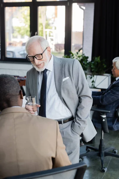 Uomo d'affari anziano sorridente che tiene un bicchiere d'acqua mentre parla con un collega afroamericano — Foto stock