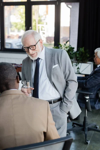 Sonriente empresario senior sosteniendo un vaso de agua mientras habla con un colega afroamericano - foto de stock