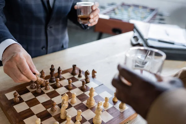 Vista parcial de hombres de negocios mayores borrosos con vasos de whisky jugando al ajedrez en la oficina - foto de stock