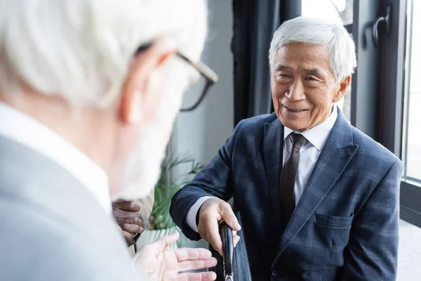 Senior asian businessman smiling while giving briefcase to blurred business partner — Stock Photo