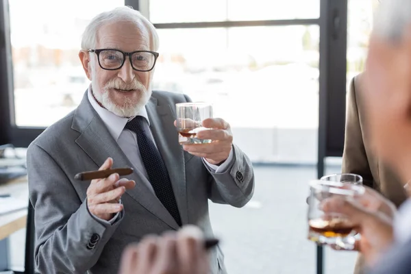 Hombre de negocios senior con vaso de whisky y cigarro cerca de colegas borrosos en la oficina - foto de stock
