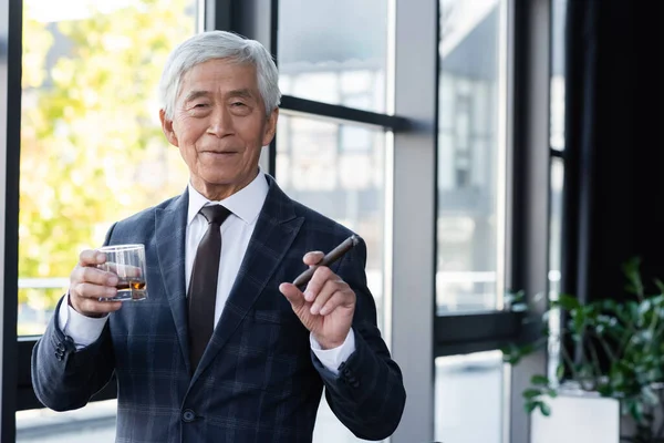 Senior asiático hombre de negocios con cigarro y vaso de whisky sonriendo a cámara en oficina - foto de stock