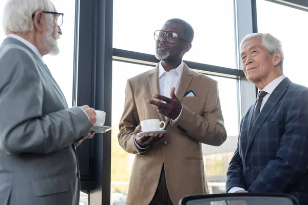 Homme d'affaires afro-américain dans les lunettes geste tout en parlant à des collègues interraciaux sur la pause café — Photo de stock