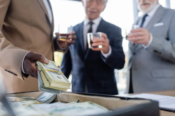 Visão recortada do empresário afro-americano segurando dólares perto da pasta e colegas seniores borrados com copos de uísque — Fotografia de Stock