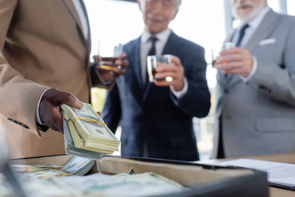 Cropped view of african american businessman holding dollars near briefcase and blurred senior colleagues with glasses of whiskey — Stock Photo