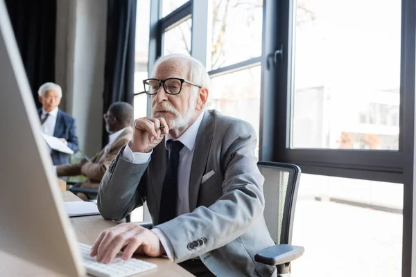 Hombre de negocios senior en gafas que trabajan en la computadora cerca de colegas difuminados multiculturales - foto de stock