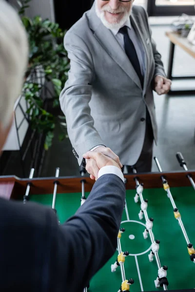 Vista cortada de empresário sênior apertando as mãos com colega borrado perto de futebol de mesa — Fotografia de Stock