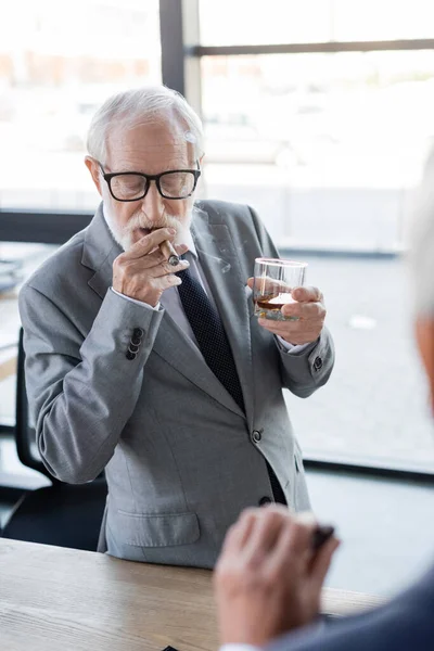 Elderly businessman in eyeglasses holding glass of whiskey while smoking cigar near blurred colleague — Stock Photo