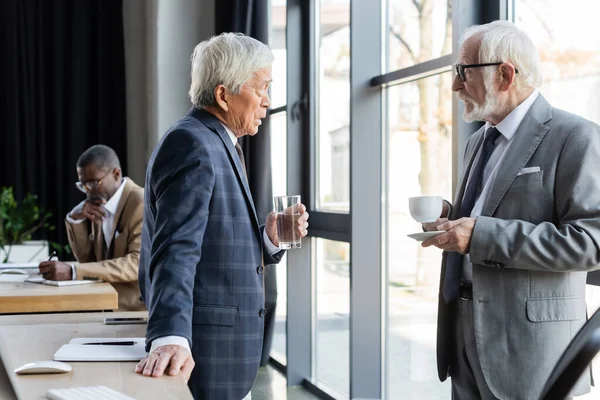 Interracial businessmen talking in office near blurred african american colleague — Stock Photo