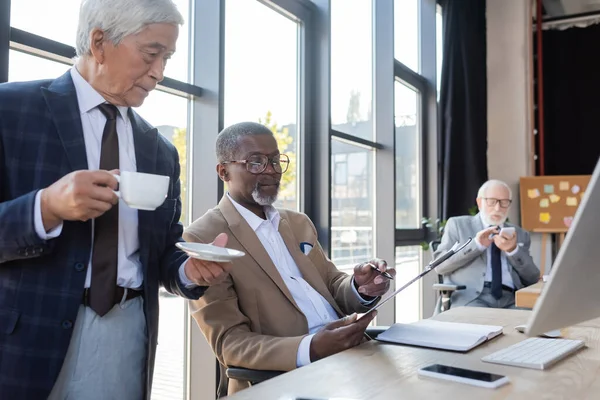 Africano americano hombre de negocios mostrando documento a senior asiático colega bebiendo café en oficina - foto de stock
