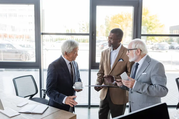 Hombres de negocios multiétnicos senior con portapapeles, taza de café y teléfono inteligente hablando en la oficina - foto de stock
