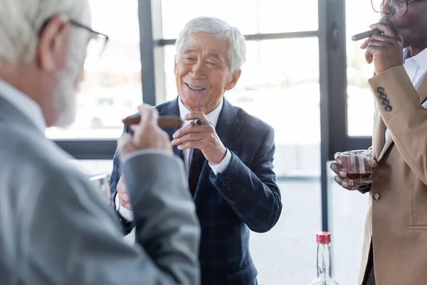 Älterer asiatischer Geschäftsmann lächelt neben Kollegen, die im Büro Zigarren rauchen und Whiskey trinken — Stockfoto