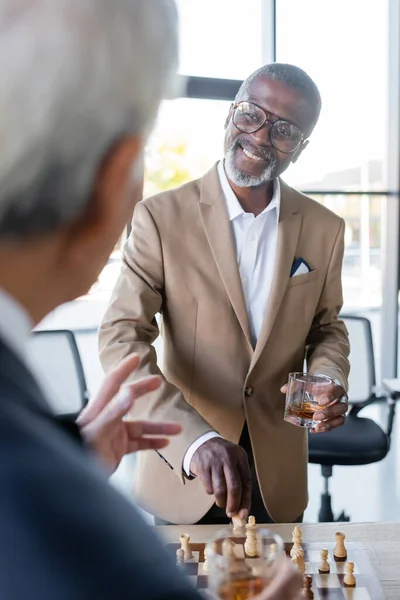 Alegre hombre de negocios afroamericano con vaso de whisky jugando al ajedrez con un colega borroso en la oficina - foto de stock